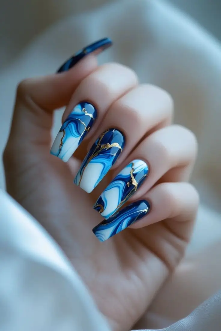 Close-up of a hand with long coffin nails featuring a blue opal marble nail art design with elegant swirls of deep blue, light blue, and white, intricately accented with gold veins, showcasing a luxurious and trending US manicure.