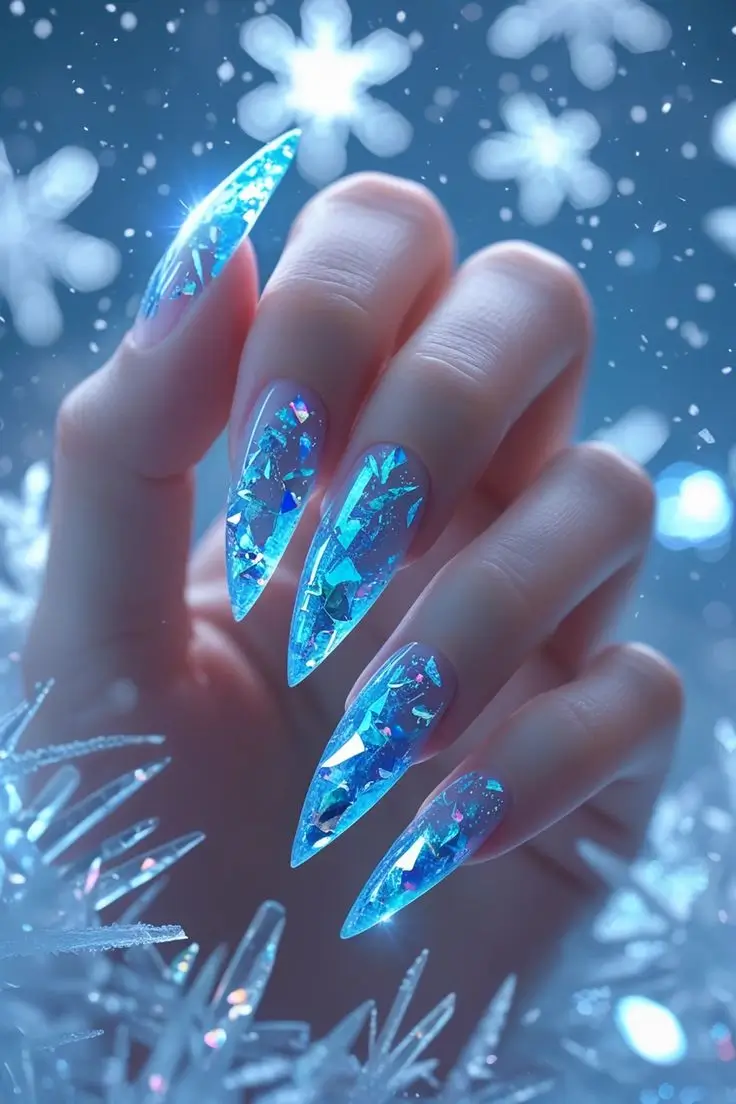 Close-up of a hand with long stiletto nails featuring crystal ice shard nail art design with clear blue tones and encapsulated holographic foil flakes for a dazzling and trending winter manicure in the USA.