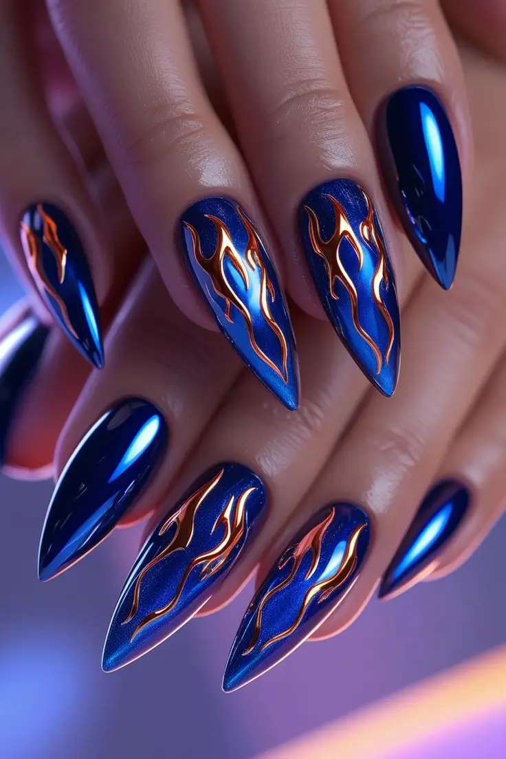 Close-up of a hand with long stiletto nails featuring an electric indigo blue chrome finish with vibrant orange and gold flame nail art, showcasing a bold and trending look for US manicures.