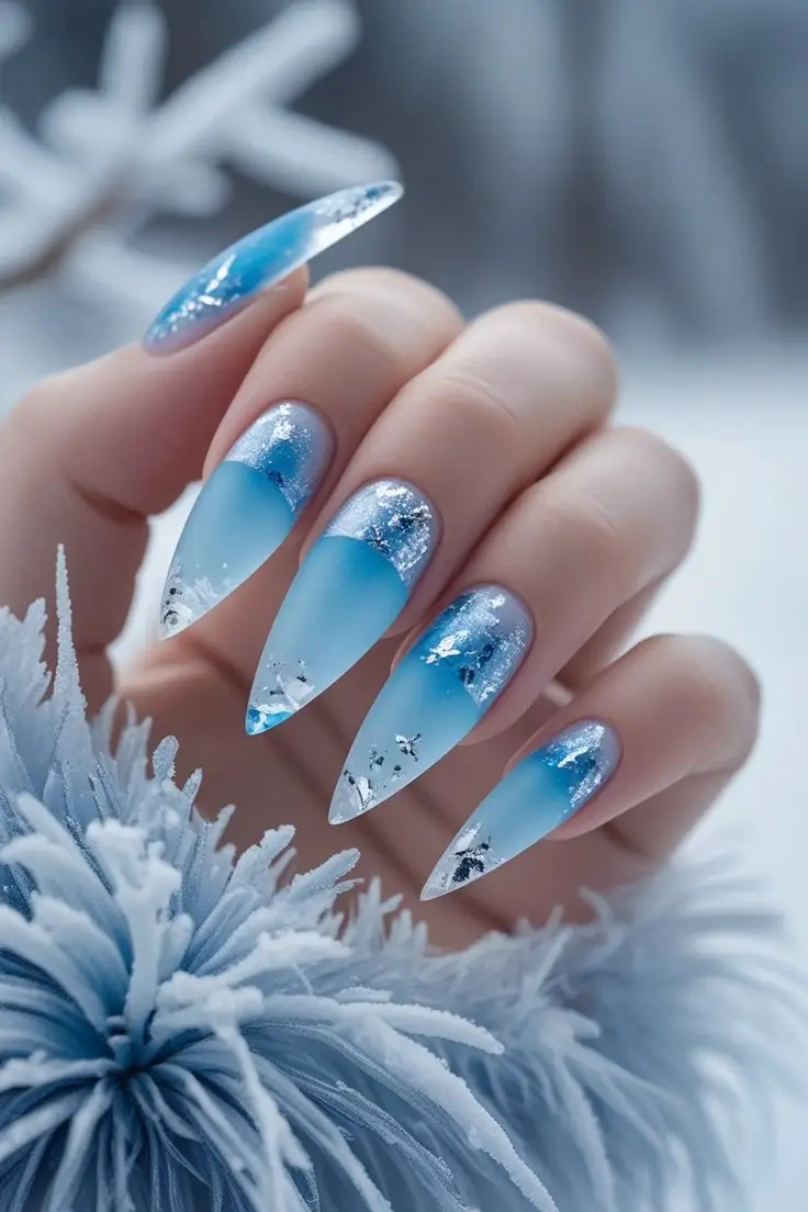 Close-up of a hand with long stiletto nails featuring a frosted glacier ice nail art design with a matte blue ombre fading to clear tips, adorned with shimmering silver flakes, representing a winter trend in USA manicures.
