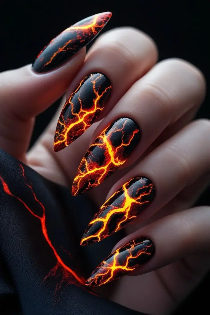 Close-up of a hand with long black stiletto nails featuring a detailed molten lava cracking nail art design in vibrant orange and red, showcasing a fierce and trending look for US nail enthusiasts.