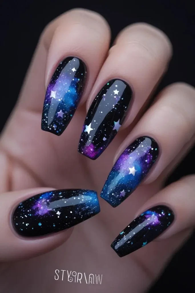 Close-up of a hand with long coffin nails featuring black galaxy glitter nail art with blue and purple nebula swirls and white stars, showcasing a cosmic and trending design for US manicures.