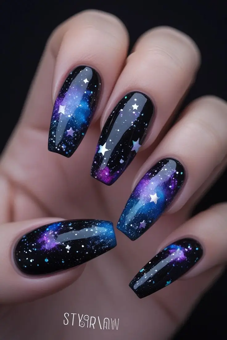 Close-up of a hand with long coffin nails featuring black galaxy glitter nail art with blue and purple nebula swirls and white stars, showcasing a cosmic and trending design for US manicures.