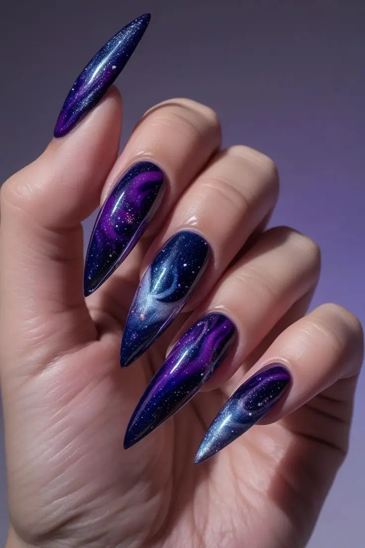 Close-up of a hand with long stiletto nails featuring molten metallic galaxy nail art with swirling blue and purple patterns and glitter, showcasing a cosmic and trending design for US manicures.