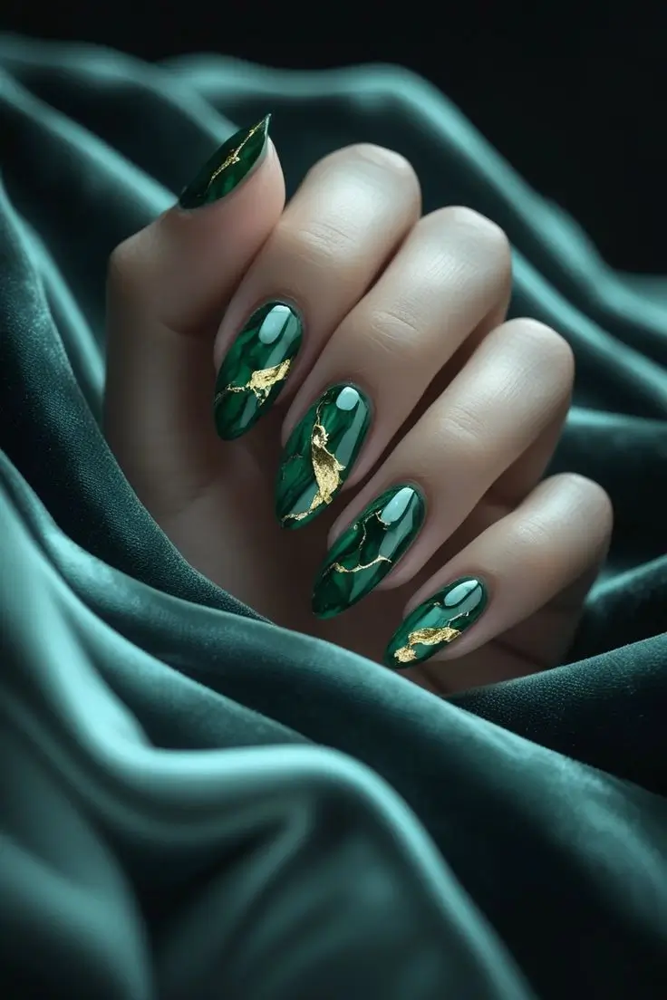 Close-up of a hand with long almond nails featuring emerald gold leaf marble nail art with a glossy deep green base, lighter green swirls, and gold foil veins, showcasing a luxurious and trending design for US manicures.