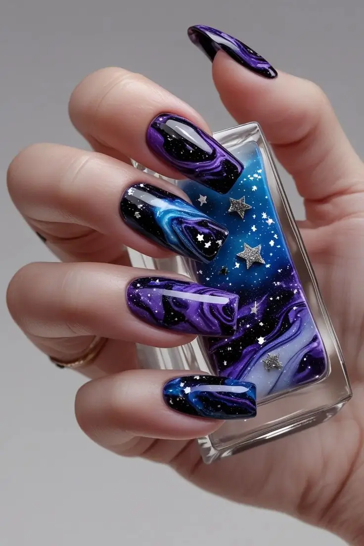 Close-up of a hand with long square nails featuring galactic marble glitter nail art with swirling purple, blue, and black cosmic patterns, silver glitter, and star foils, showcasing a mystical and trending design for US manicures.