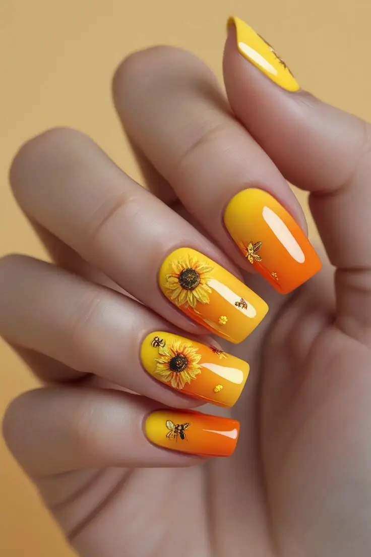 Close-up of a hand with coffin nails featuring a sunflower honey ombre nail design with a yellow to orange gradient, hand-painted sunflowers, and bee decals, showcasing a summer-inspired and trending design for US manicures.